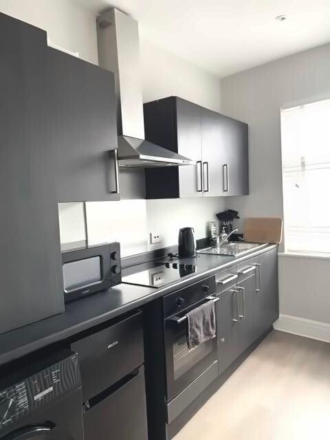 A modern kitchen features sleek black cabinetry and stainless steel appliances, including a hob, oven, and microwave. A kettle and cutting board are placed on the countertop. Natural light filters in through a nearby window, creating a bright cooking space.