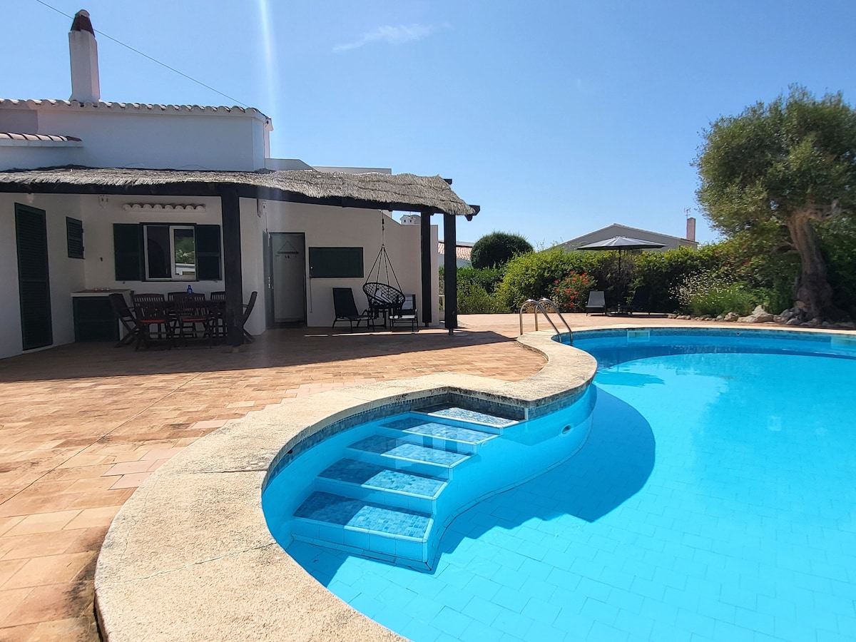 An inviting outdoor area features a spacious pool with steps leading into the water. A shaded terrace provides seating and dining options, surrounded by lush greenery. Nearby, lounge chairs offer a place to relax under clear skies.