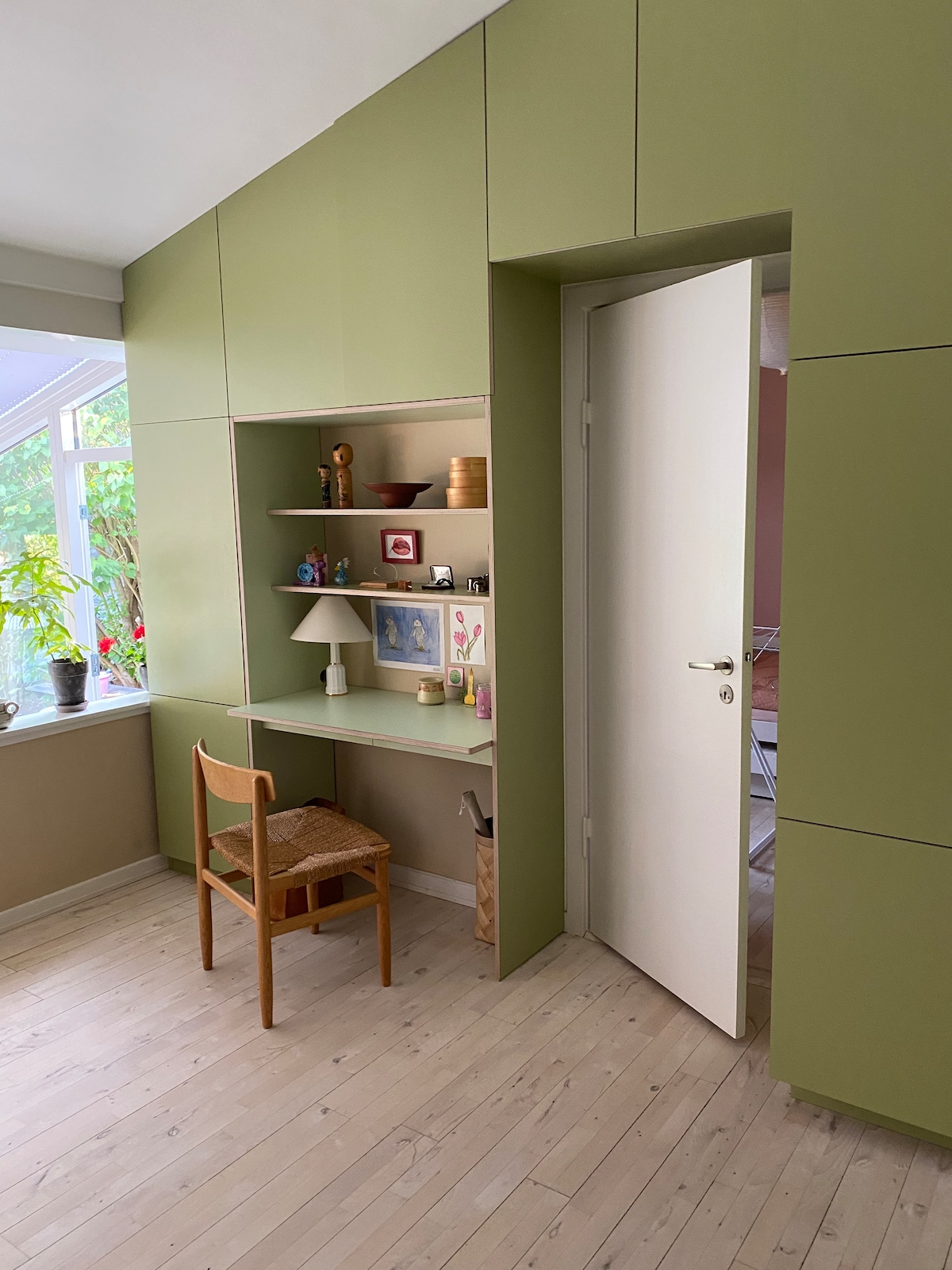 A compact workspace is integrated into a wall unit featuring light green cabinetry. A wooden chair is positioned at a small desk, adorned with personal items, and a lamp. A window allows natural light to illuminate the area, with indoor plants adding a touch of greenery.