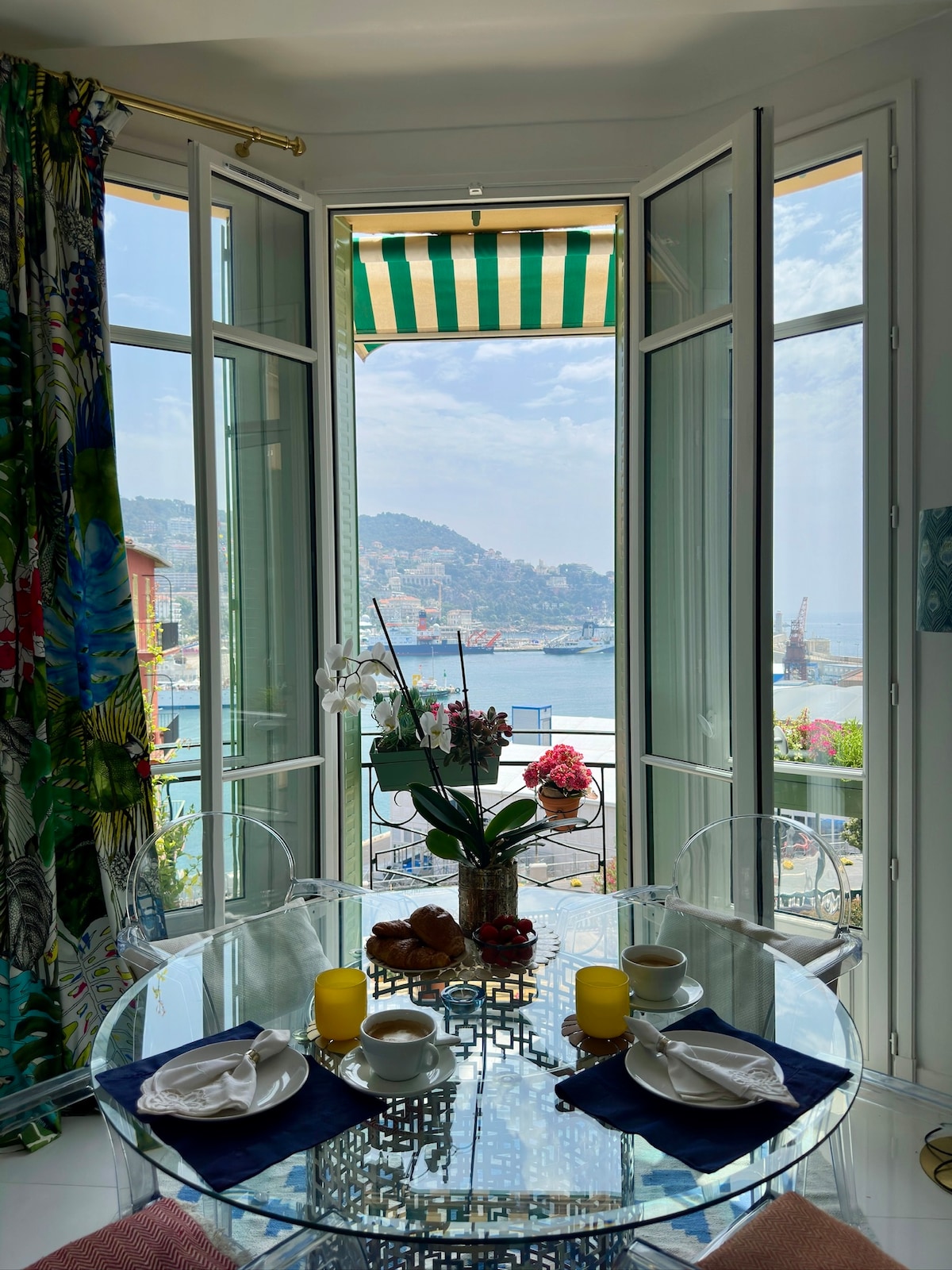 A bright dining area features a round glass table set with tableware, surrounded by clear chairs. Large windows open to reveal a spectacular sea view, framed by colorful curtains. Fresh flowers and pastries add to the inviting setting, creating a serene space for meals.
