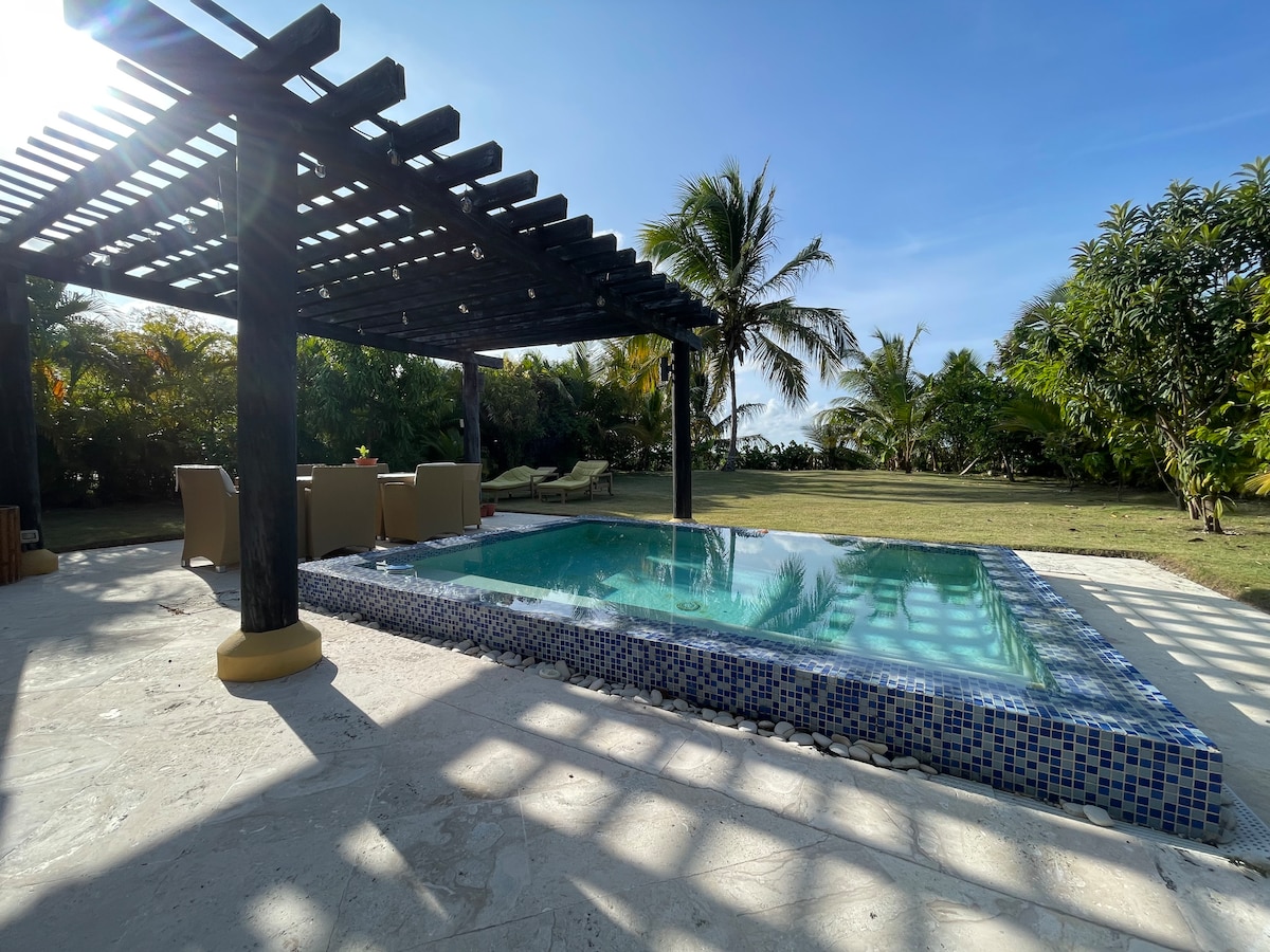 A private pool is set within a serene outdoor area, surrounded by lush greenery and palm trees. A shaded seating area featuring chairs is positioned beside the pool, offering a perfect spot for relaxation. Sunlight filters through the overhead structure, casting playful shadows on the ground.
