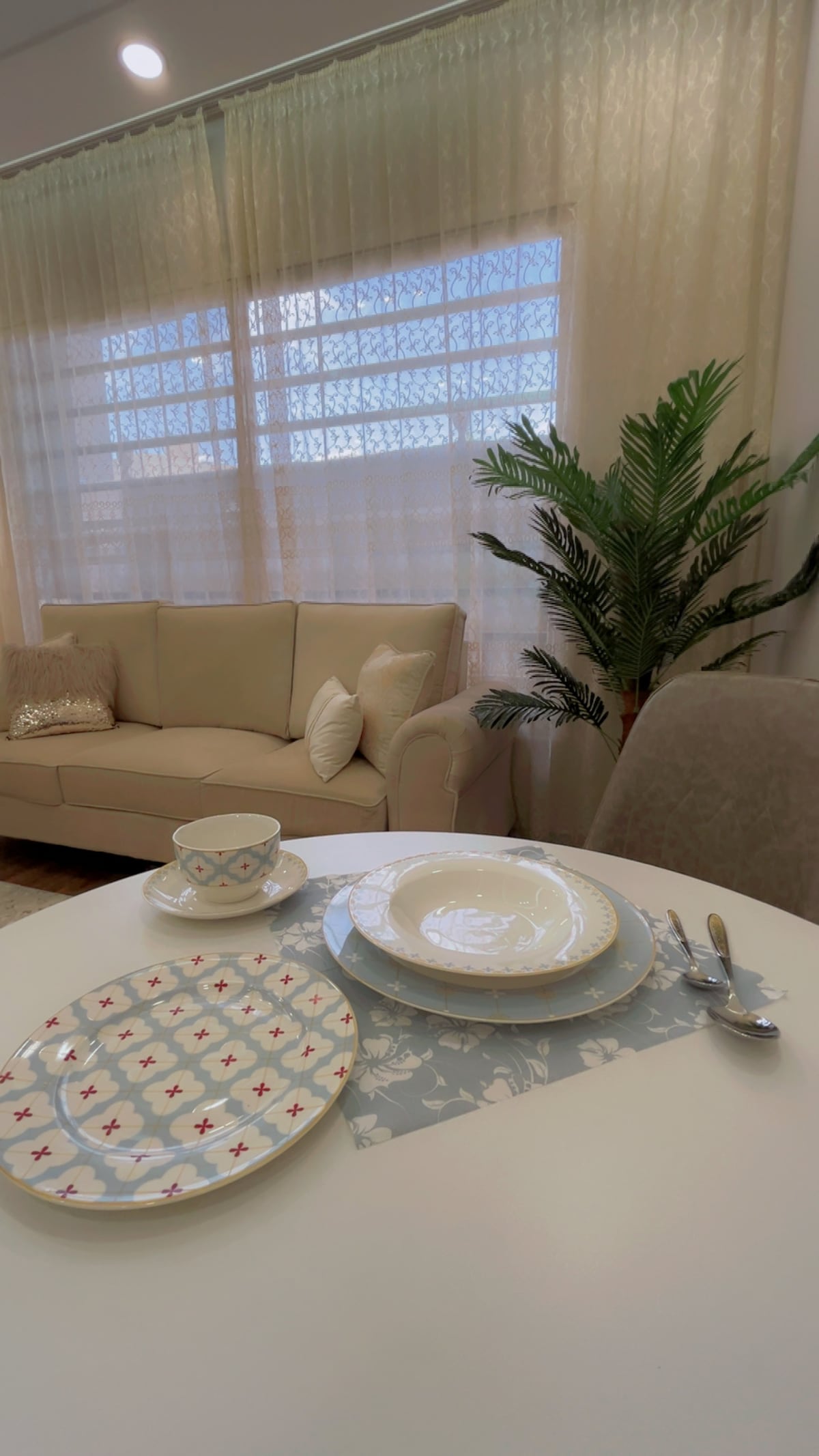 A dining table is set with plates and utensils, showcasing a decorative plate featuring a star pattern. A light-colored sofa is visible in the background, complemented by soft cushions and a potted plant beside it, while natural light filters through sheer curtains.