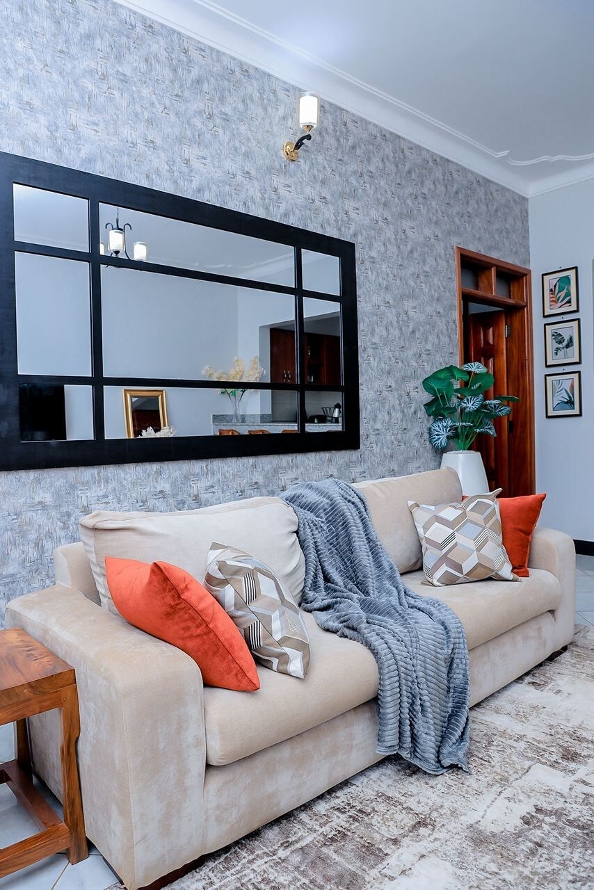 A comfortable seating area features a light-colored sofa adorned with decorative pillows in earthy tones. A soft throw blanket drapes over one side. A large mirror is positioned on the wall, reflecting the adjacent room, while framed art adds character to the space.