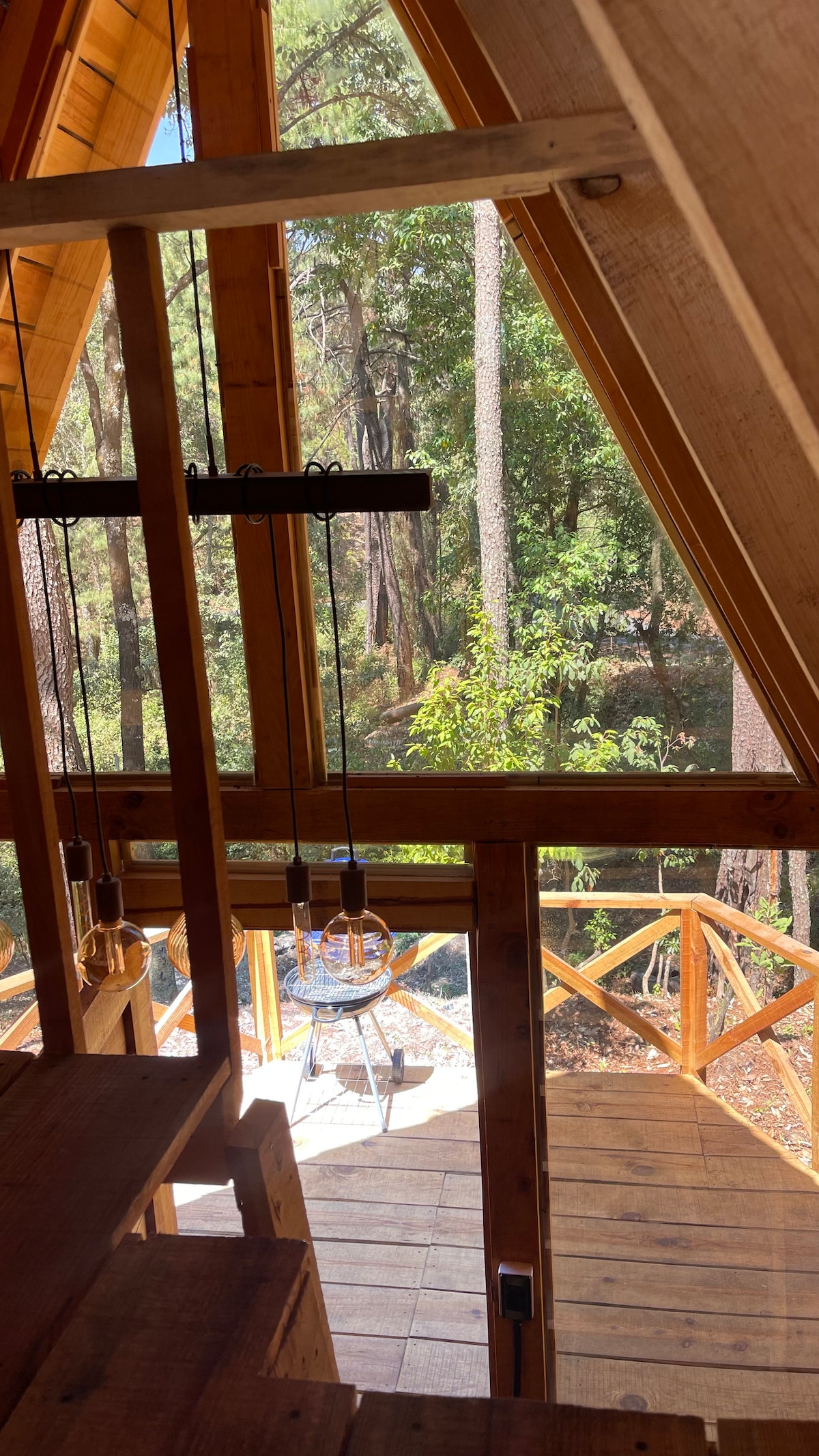 Natural light floods through large A-frame windows, illuminating wooden beams and stairs. A view of a deck with outdoor seating reveals a backdrop of lush trees and foliage, enhancing the connection to the surrounding nature.