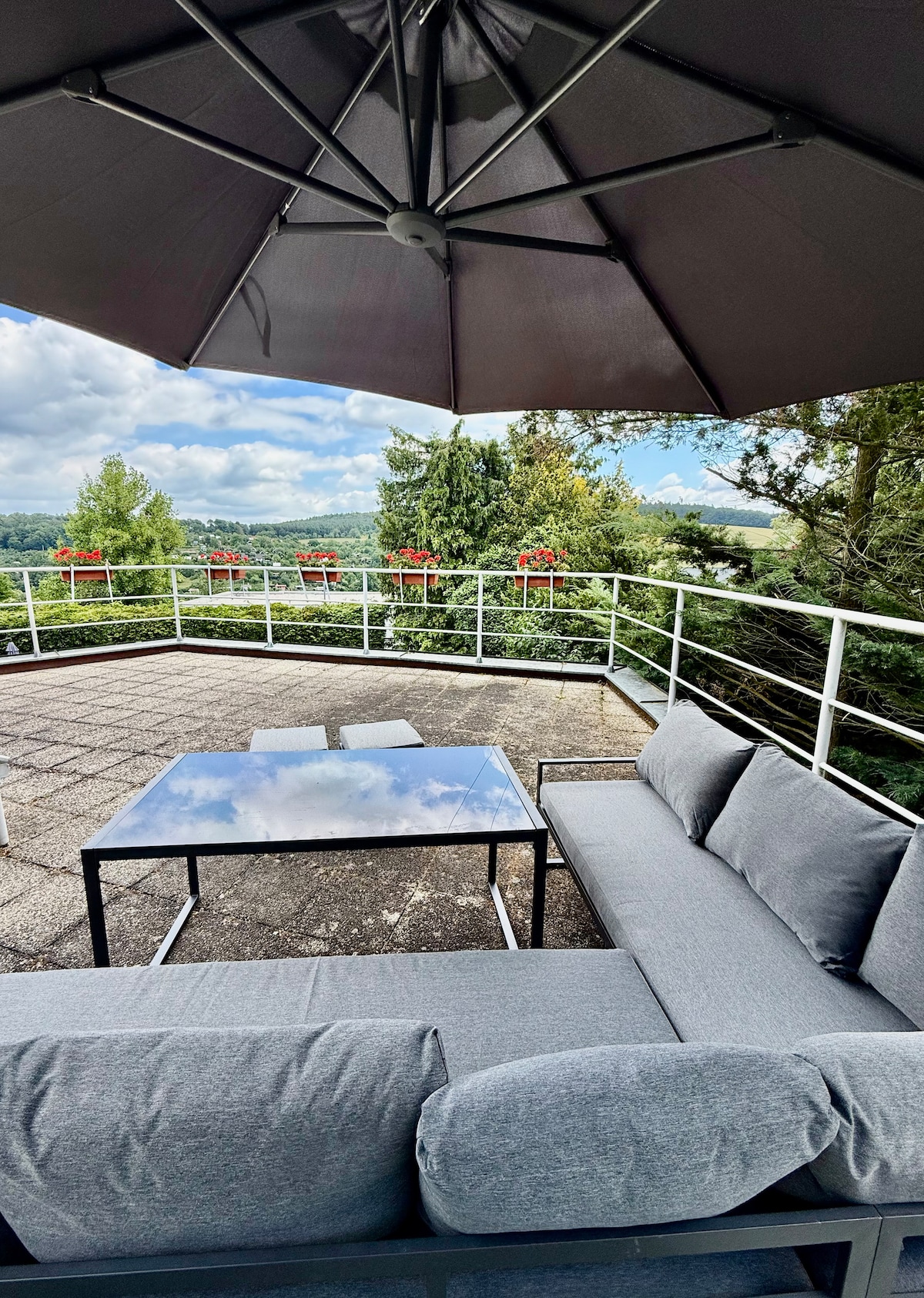 A comfortable outdoor seating area features a gray cushioned sofa and a glass-top table, positioned under a large umbrella. A railing offers a view of surrounding greenery, while clouds create a serene sky in the background.