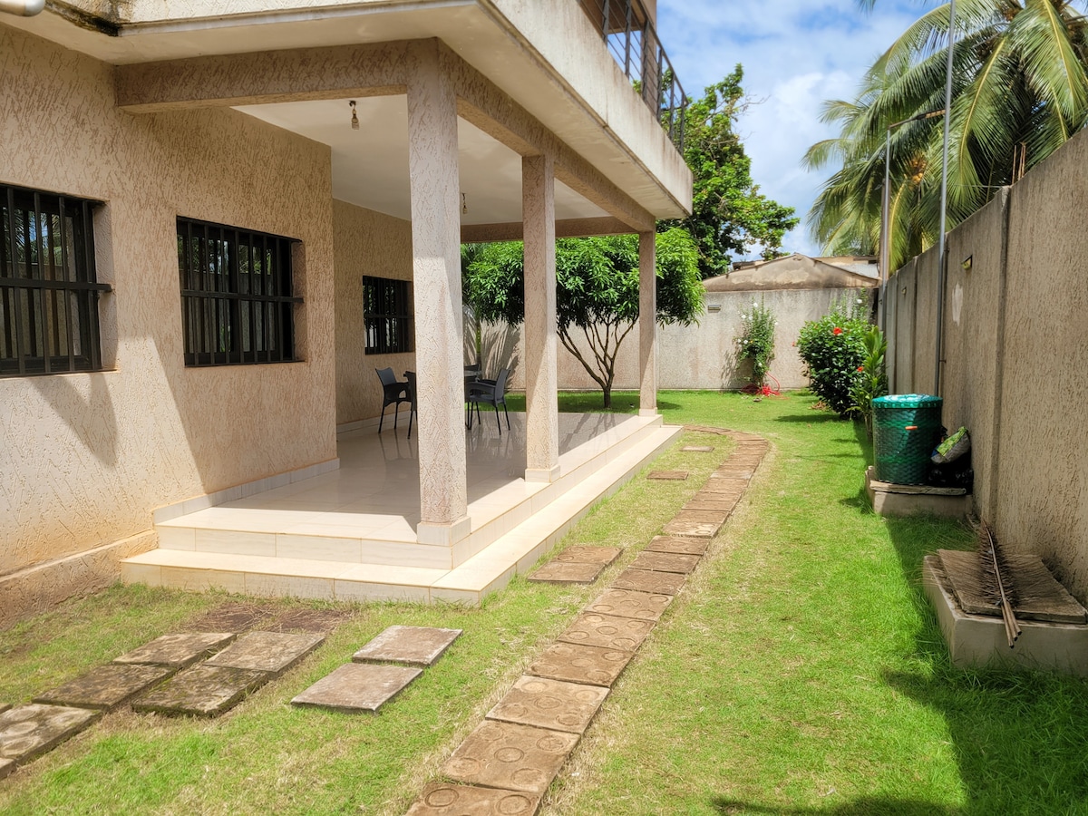 A spacious outdoor area features a patio with four chairs and a table, positioned under a covered verandah. A well-maintained garden is outlined by stone pathways, surrounded by greenery and several trees, contributing to a relaxed outdoor environment.