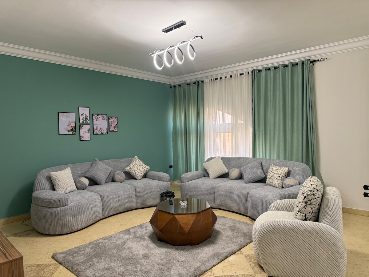 A comfortable seating area is presented with two spacious, curved sofas adorned with patterned cushions. A cozy area rug lies beneath a modern coffee table, featuring a geometric design. Light-filtering curtains frame the windows, and decorative wall art adds a subtle touch.
