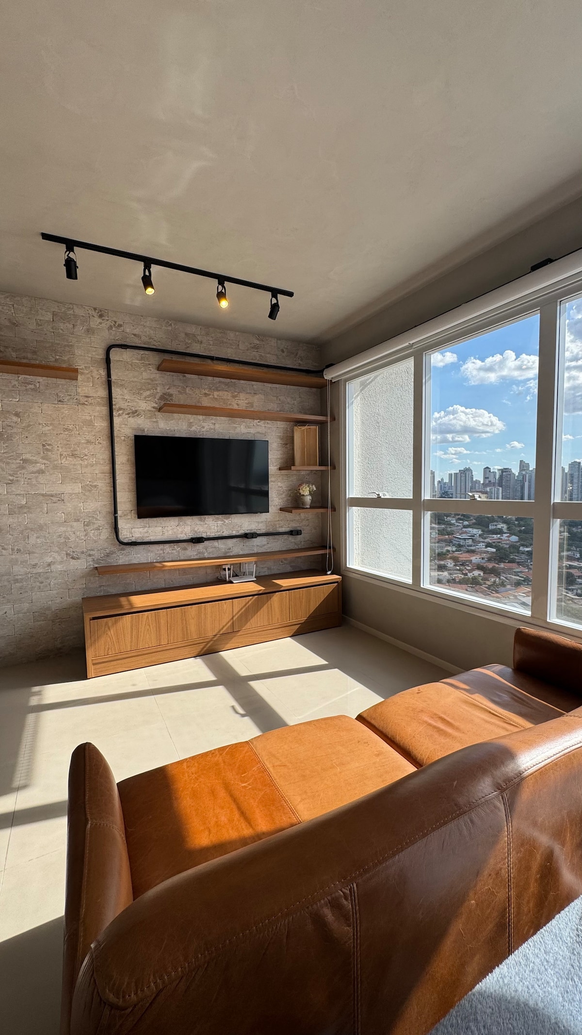 A bright living area features a comfortable brown leather sofa facing a mounted flat-screen television. Natural light floods the room through large windows that offer a view of the city skyline. Subtle shelving adorns the wall, enhancing the modern decor.