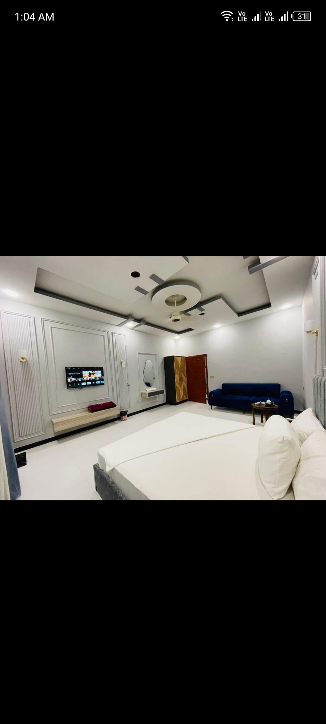 A spacious room features a modern design with a light-colored bed and multiple decorative elements on the walls. A centrally placed ceiling fan adds functionality, while a flat-screen TV is mounted on one wall. A dark sofa and small table are arranged in the corner.