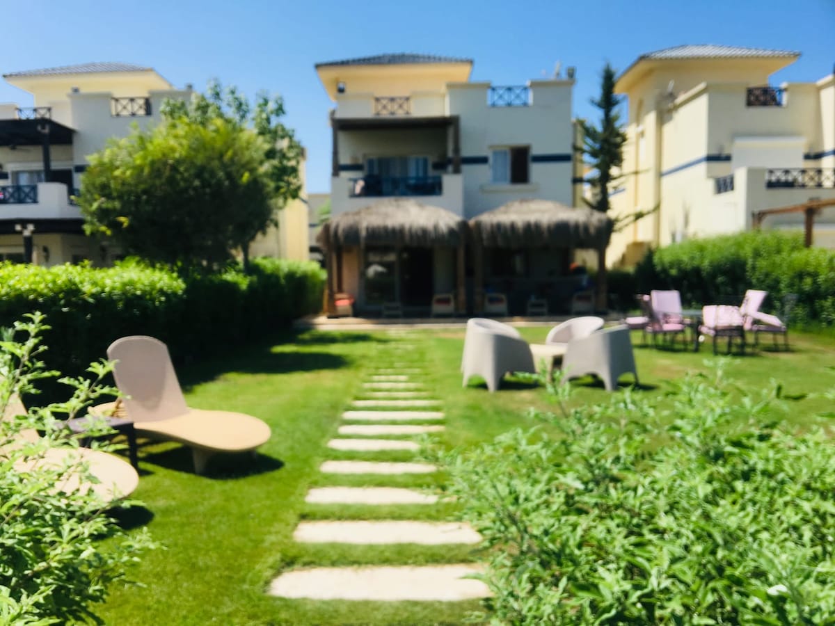A landscaped garden area is seen, featuring a stone pathway leading to a shaded seating space adorned with thatched roofs. A mix of lounge chairs and outdoor tables are arranged amidst lush greenery, inviting moments of relaxation in a tranquil setting.