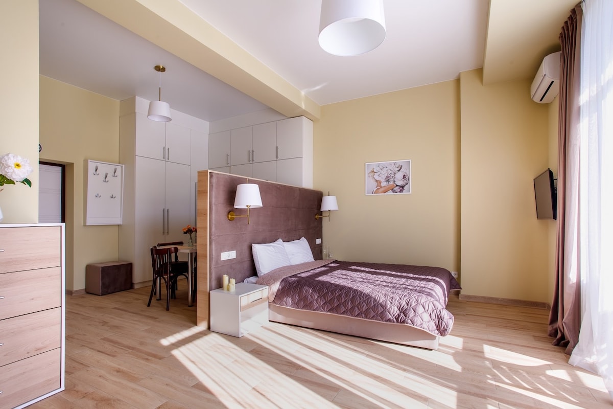 A spacious room features light yellow walls and natural wood flooring. A large bed with a textured brown headboard is positioned near a window. A contemporary dresser and a chair are visible, alongside a wall-mounted television. Soft light filters through sheer curtains.