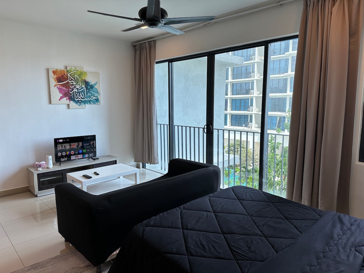 A modern room features a spacious bed with a dark comforter and a sleek black sofa. Large glass doors allow natural light to fill the space, revealing a balcony view of a pool and surrounding greenery. A flat-screen television is positioned on a white coffee table.
