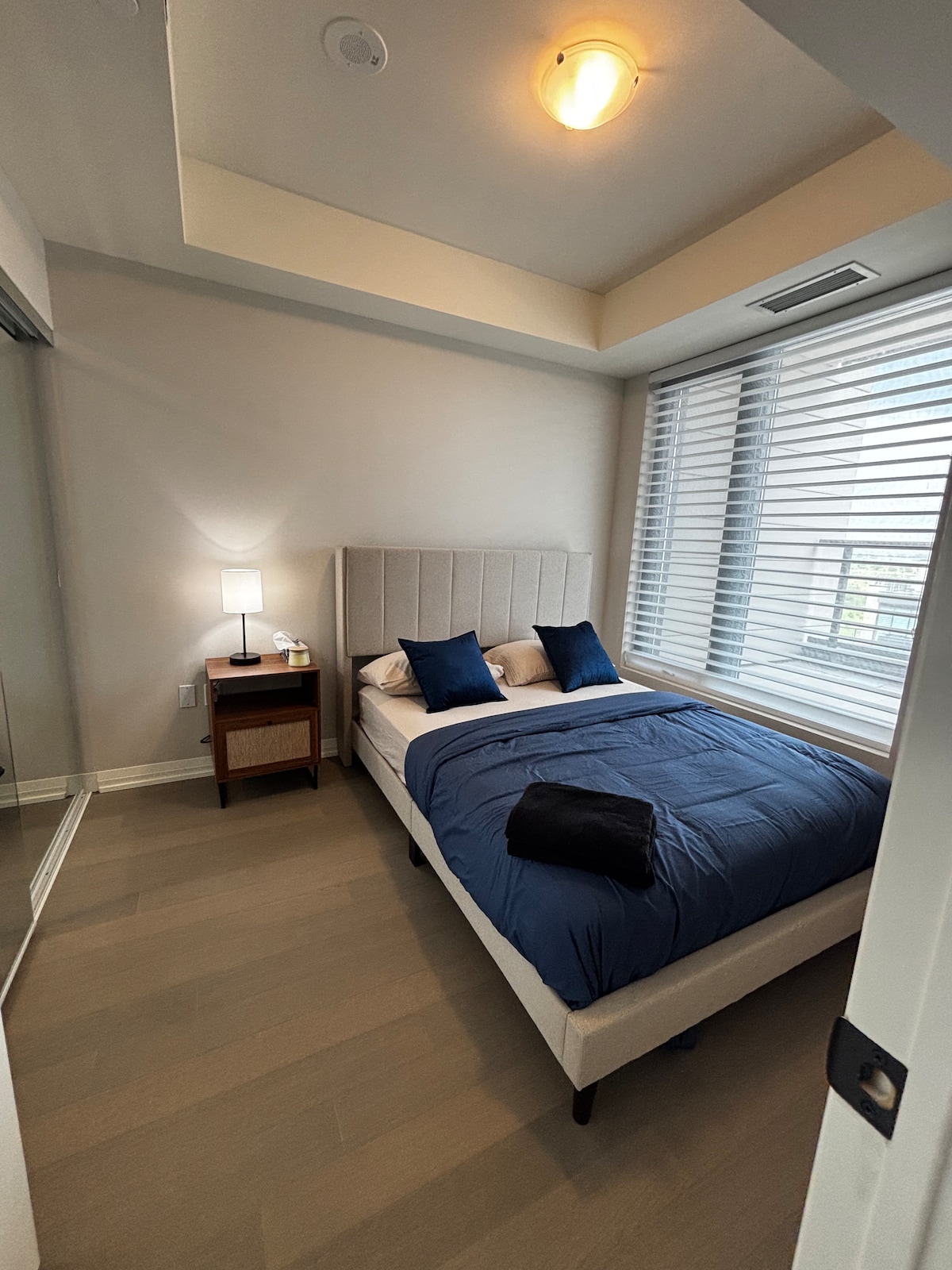 A well-lit bedroom features a spacious bed dressed in blue linens, complemented by two decorative pillows. A bedside table with a lamp provides soft illumination. Large windows with horizontal blinds allow natural light to filter in, enhancing the room's modern aesthetic.