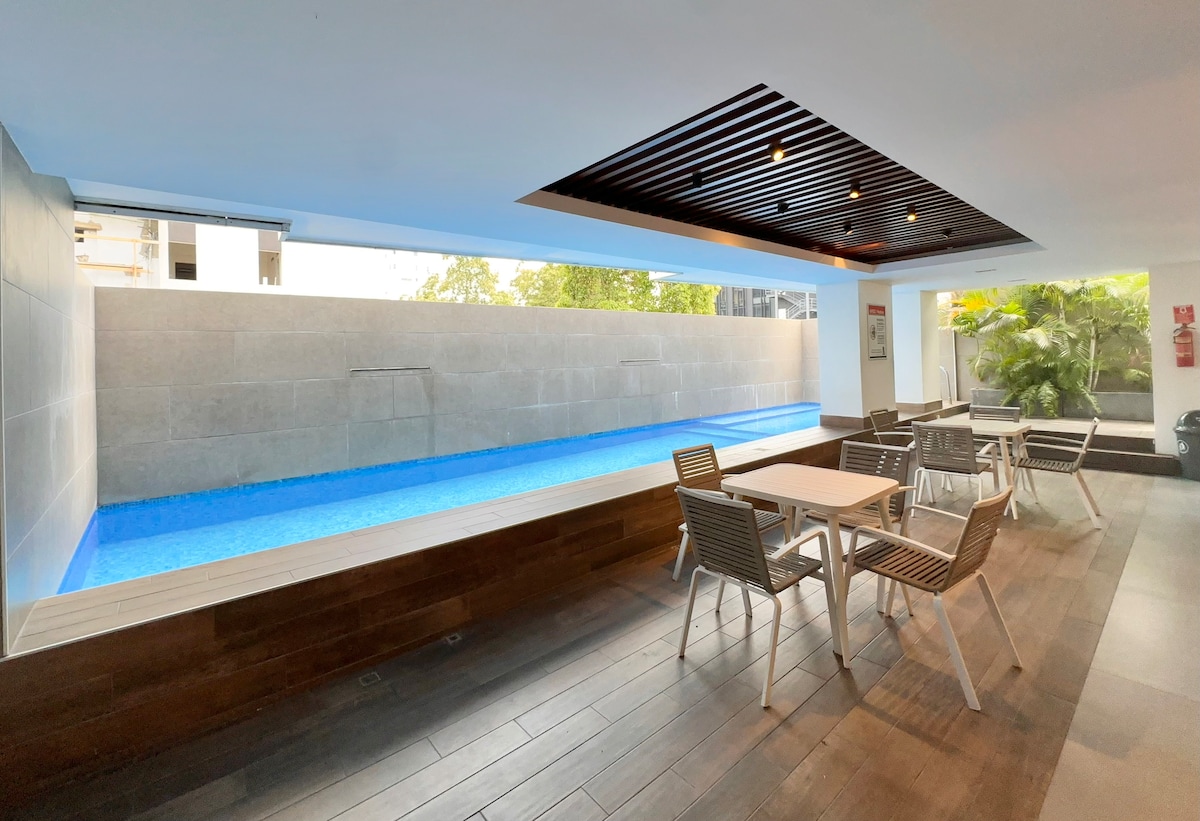 An inviting communal area features a serene pool with vibrant blue water and a sleek stone wall backdrop. Several tables and chairs are arranged on the wooden deck, providing a comfortable space for relaxation and socializing amidst lush greenery.