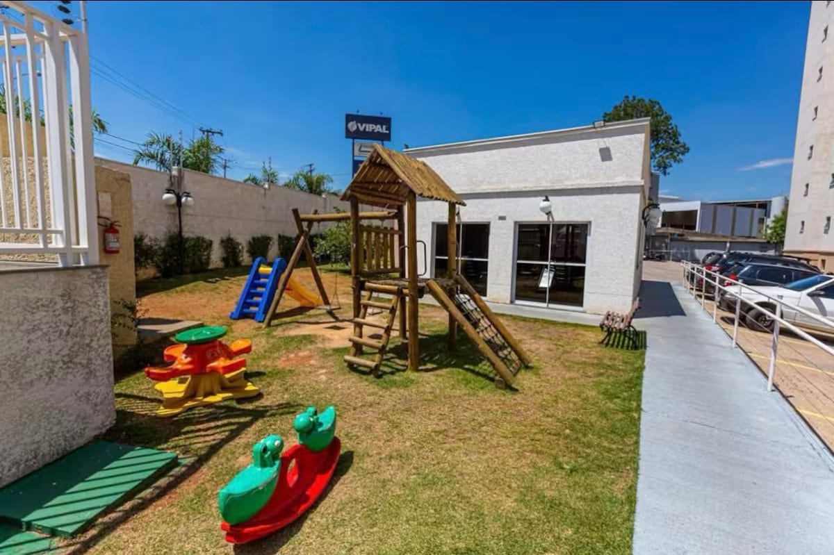 A children's play area is visible, featuring a wooden playset with slides and climbing features. Colorful plastic tables and chairs are arranged nearby. A well-maintained green lawn surrounds the play area, with clear blue skies overhead and a building with large windows in the background.