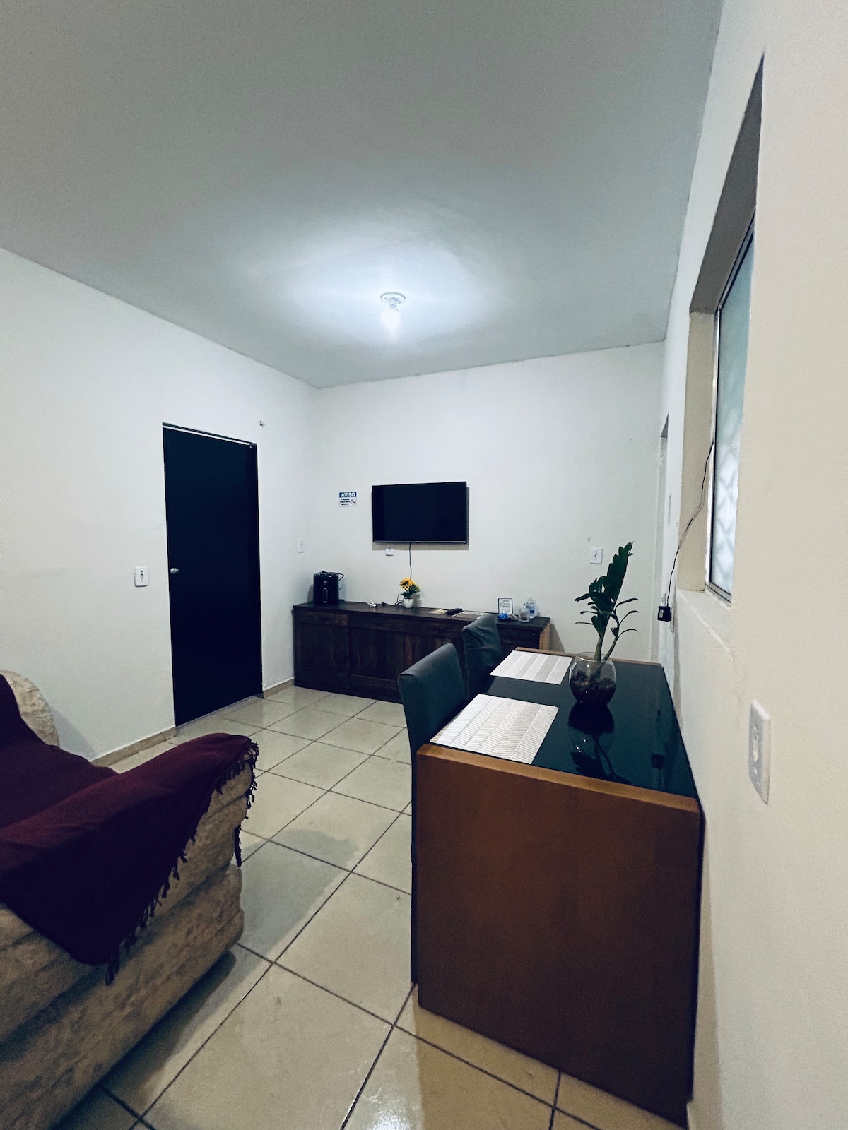 A functional living area is visible, featuring a dark wooden cabinet and a wall-mounted TV. A desk with two chairs is positioned beside a small plant, while a soft sofa adds to the space's comfort. Natural light enters through a nearby window.