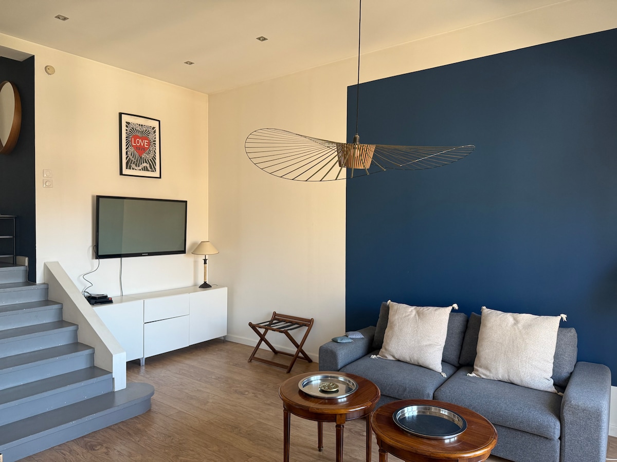 A modern living area is showcased, featuring a comfortable gray sofa with light cushions and two circular wooden side tables. A flat-screen TV is mounted on a white media console, and a unique ceiling fan adds a contemporary touch. Natural light fills the space, enhancing the neutral color palette.