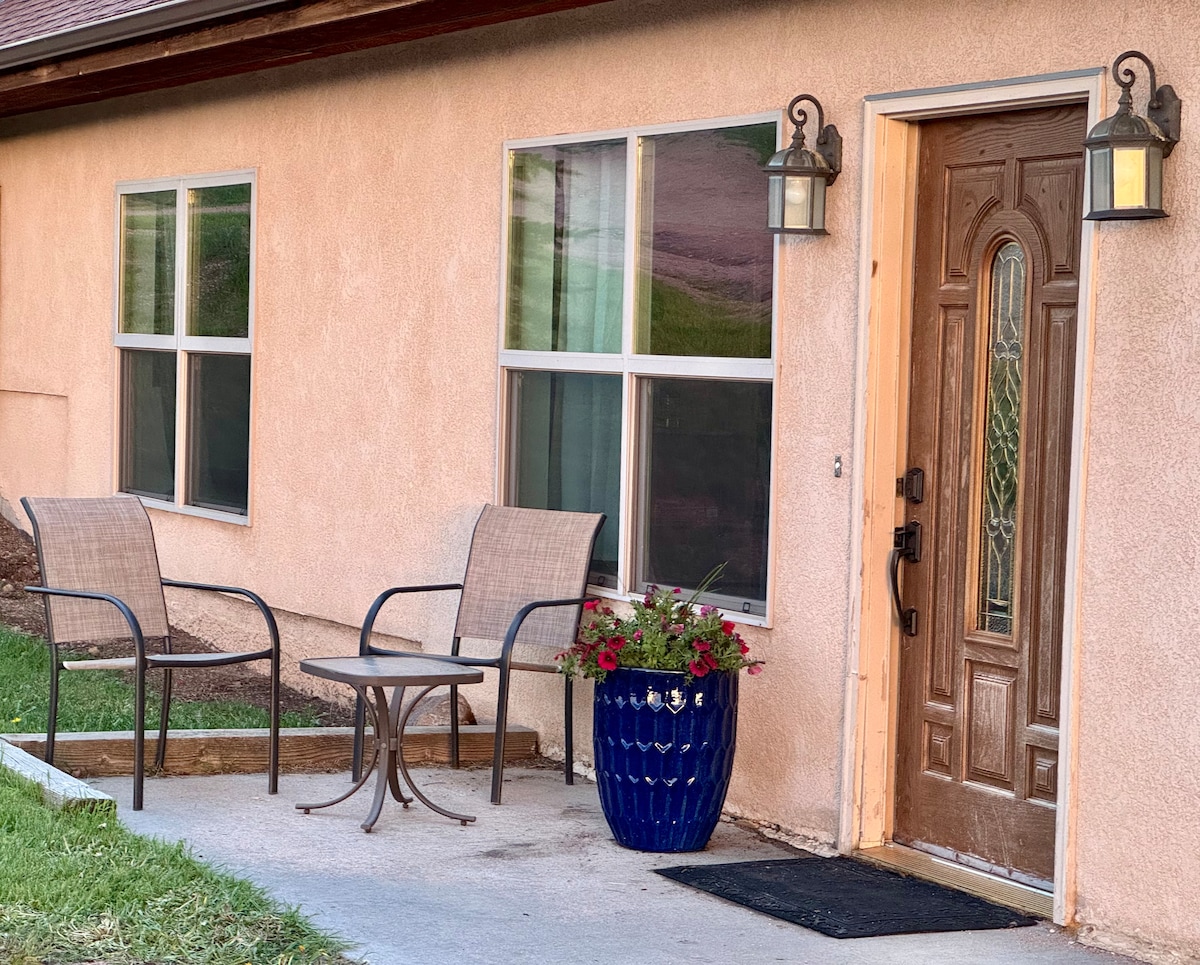 A welcoming entrance features a wooden front door with decorative glass and two light fixtures on either side. Two chairs and a small table are positioned on the patio, accompanied by a potted plant filled with colorful flowers. Large windows provide natural light.