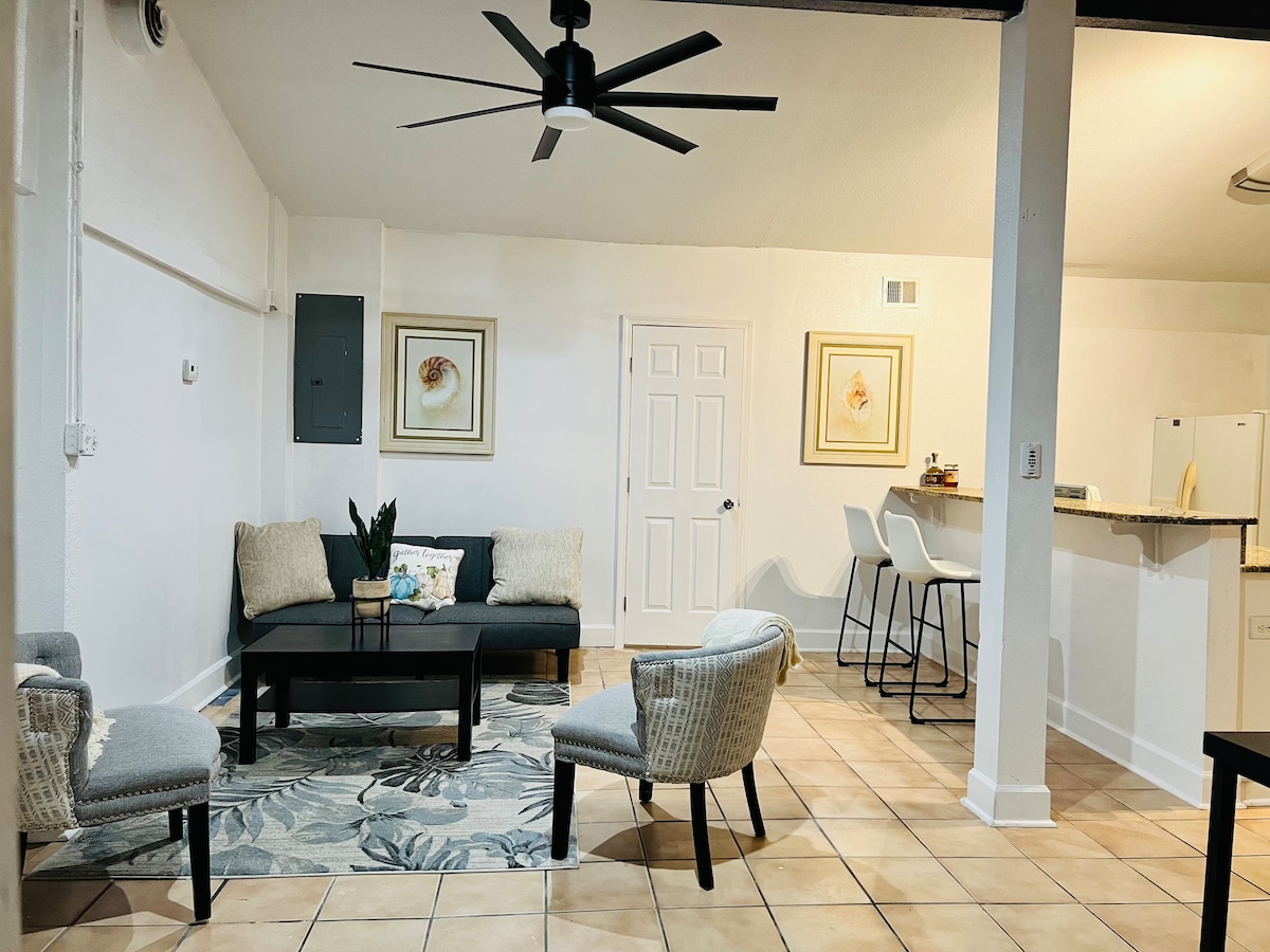 A living area features a comfortable seating arrangement with a central coffee table and decorative rug. Light-colored walls provide a bright ambiance, while a ceiling fan offers ventilation. The bar area with high stools is visible in the background, contributing to a spacious atmosphere.