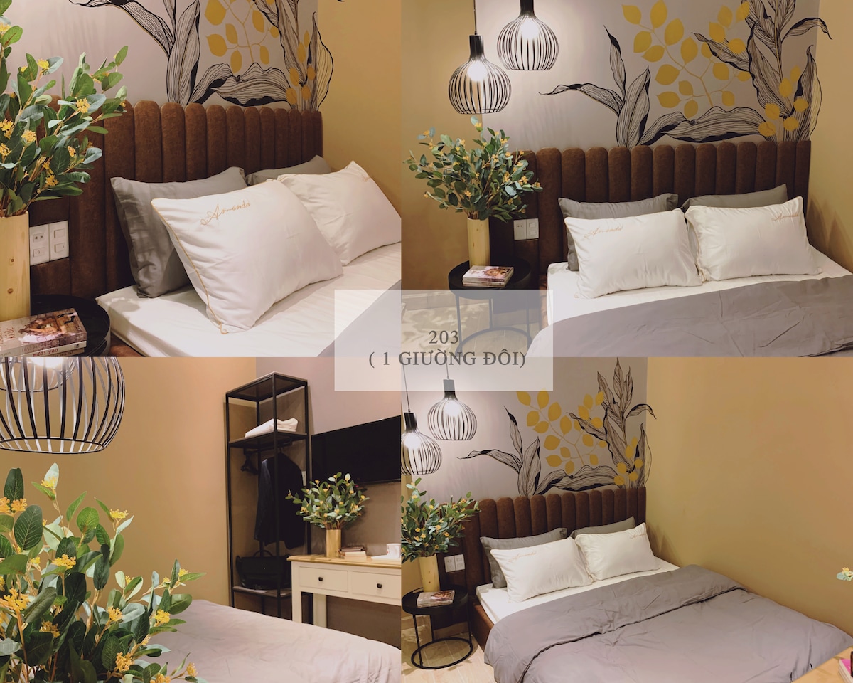 The sleeping area features a spacious bed with a tufted headboard. Soft, white pillows and a light gray blanket are arranged neatly. A decorative plant adds a touch of greenery, while a wall mural of plants enhances the ambiance. Subtle lighting is provided by hanging fixtures.