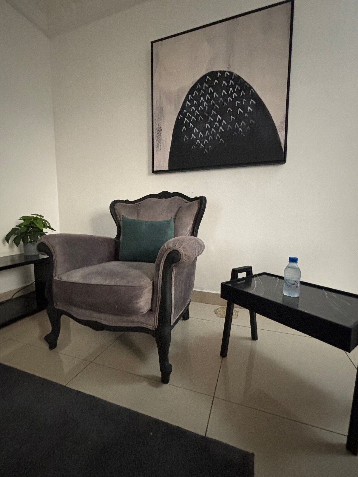 A cozy seating area features a plush, velvet armchair with ornate detailing, accompanied by a small black side table. A stylish piece of wall art depicting a mountain silhouette is displayed above the chair. A green plant brings a touch of nature to the space.