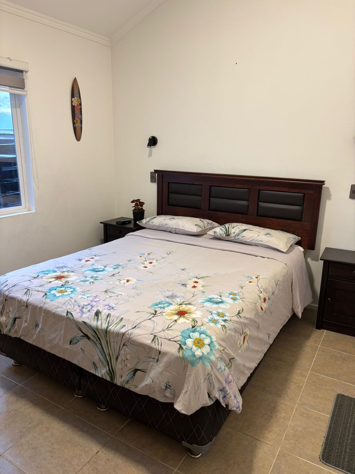 A cozy sleeping area features a double bed with a floral-patterned bedspread. Nightstands are positioned on either side of the bed, and a window allows natural light to brighten the space. A decorative wall piece adds a touch of character.