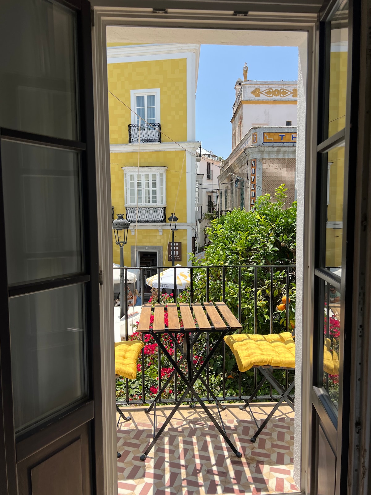 A view from open double doors reveals a small balcony with a wooden table and two yellow cushioned stools. Colorful flowers and greenery are visible in the foreground, while charming buildings in warm hues create a vibrant backdrop.