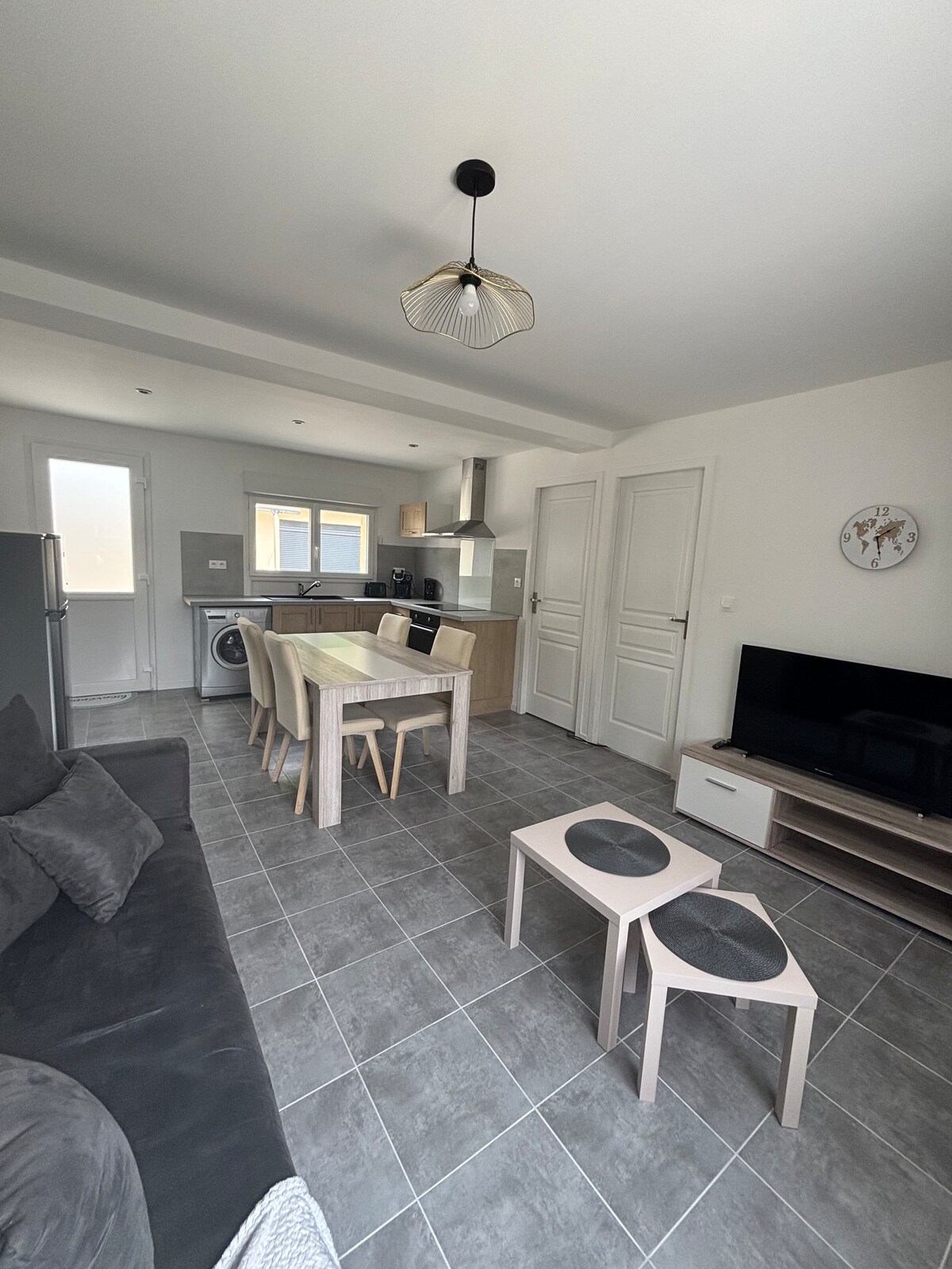 A modern living area features a cozy sofa and a wooden dining table surrounded by light-colored chairs. A sleek television sits atop a low cabinet. Large windows allow natural light to fill the space, complemented by neutral wall tones and tiled flooring.