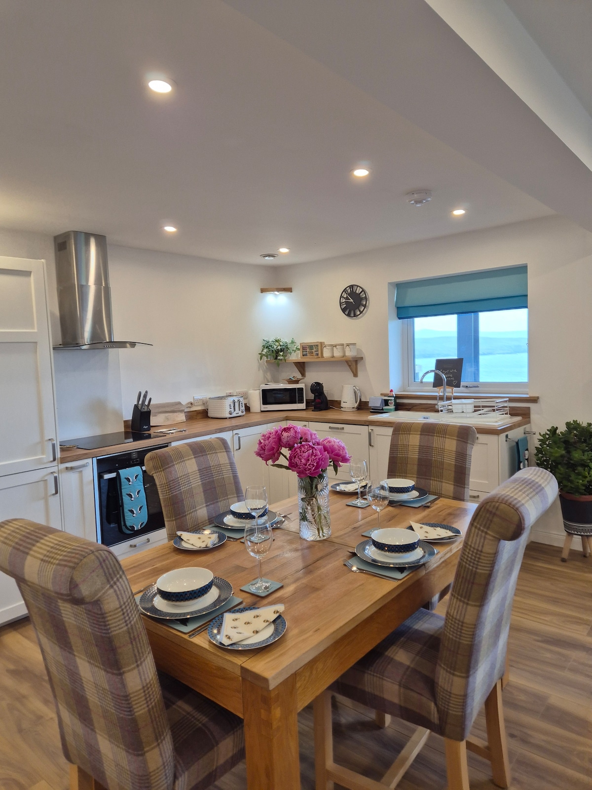 A dining table is set with plates, cutlery, and glasses, accompanied by a vase of pink flowers. The modern kitchen is visible in the background, featuring sleek appliances and a large window offering views of the sea.