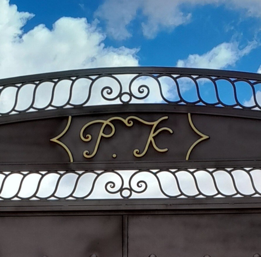 The entrance gate is adorned with elegant metalwork, featuring the letters 'P.K.' prominently displayed at the top. A backdrop of soft clouds against a blue sky adds a pleasant contrast to the gate's design.