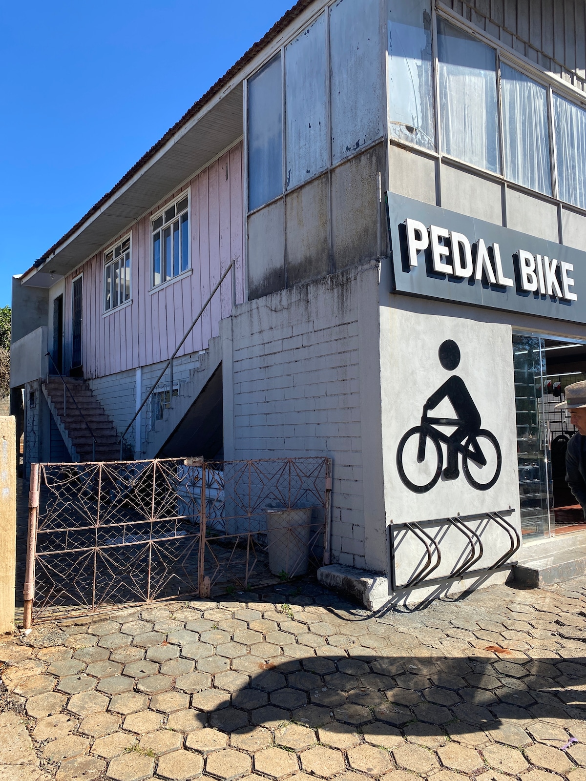 A two-story building is shown with a light pink facade and large windows. An outdoor staircase leads to the upper level, while a bike rack is positioned in front of the entrance labeled 'PEDAL BIKE.' Clear blue skies complement the scene.