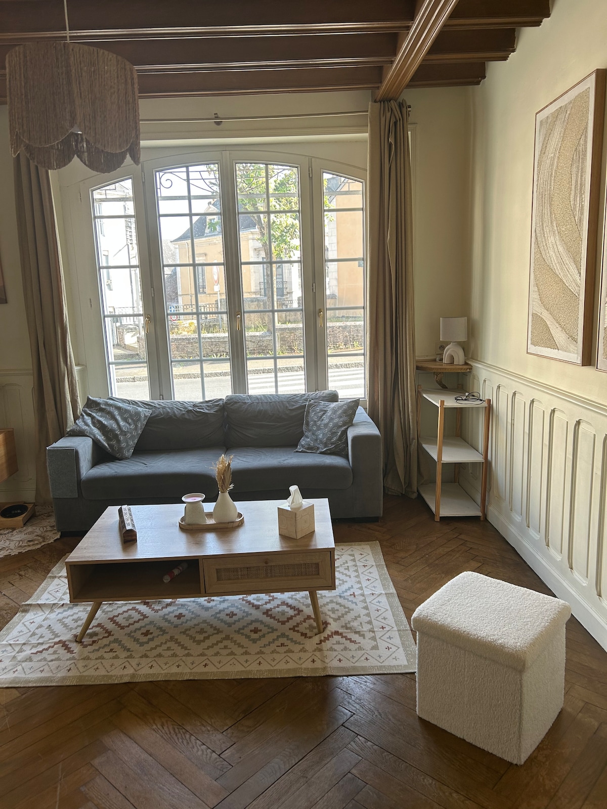 The living area features a comfortable gray sofa positioned against a light wall, complemented by a decorative rug and a wooden coffee table. Natural light enters through large glass doors, which open to a view of the outside. A small side table and a cozy ottoman are also visible.
