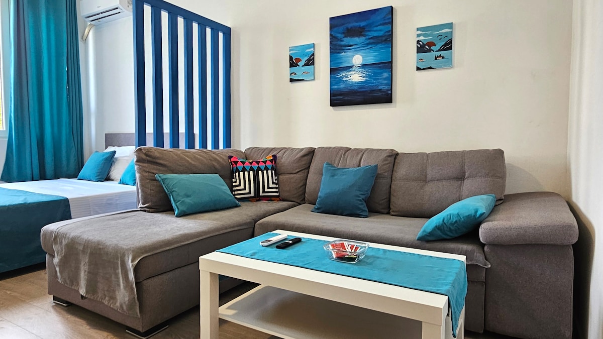 A modern studio is presented, featuring a grey sectional sofa adorned with teal accent pillows. A small white coffee table sits in front, decorated with a bowl of red items. On the wall, a vibrant blue painting hangs, complemented by two smaller artworks. Light blue drapes frame a window, enhancing the overall ambiance.