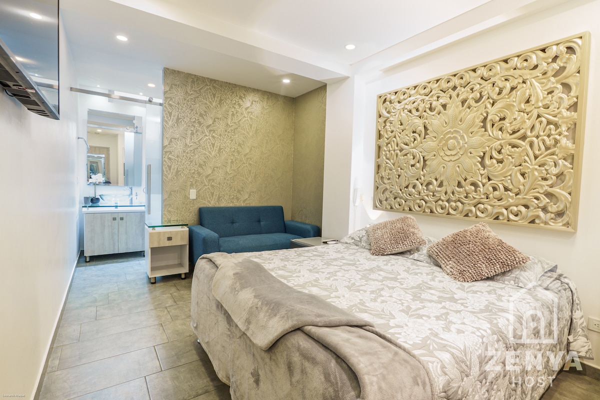 A spacious bedroom features a large bed adorned with a light-colored blanket and decorative pillows. A textured wall serves as a backdrop, while a modern sofa is positioned nearby. A well-lit bathroom area with a mirror and sink is visible in the reflection.