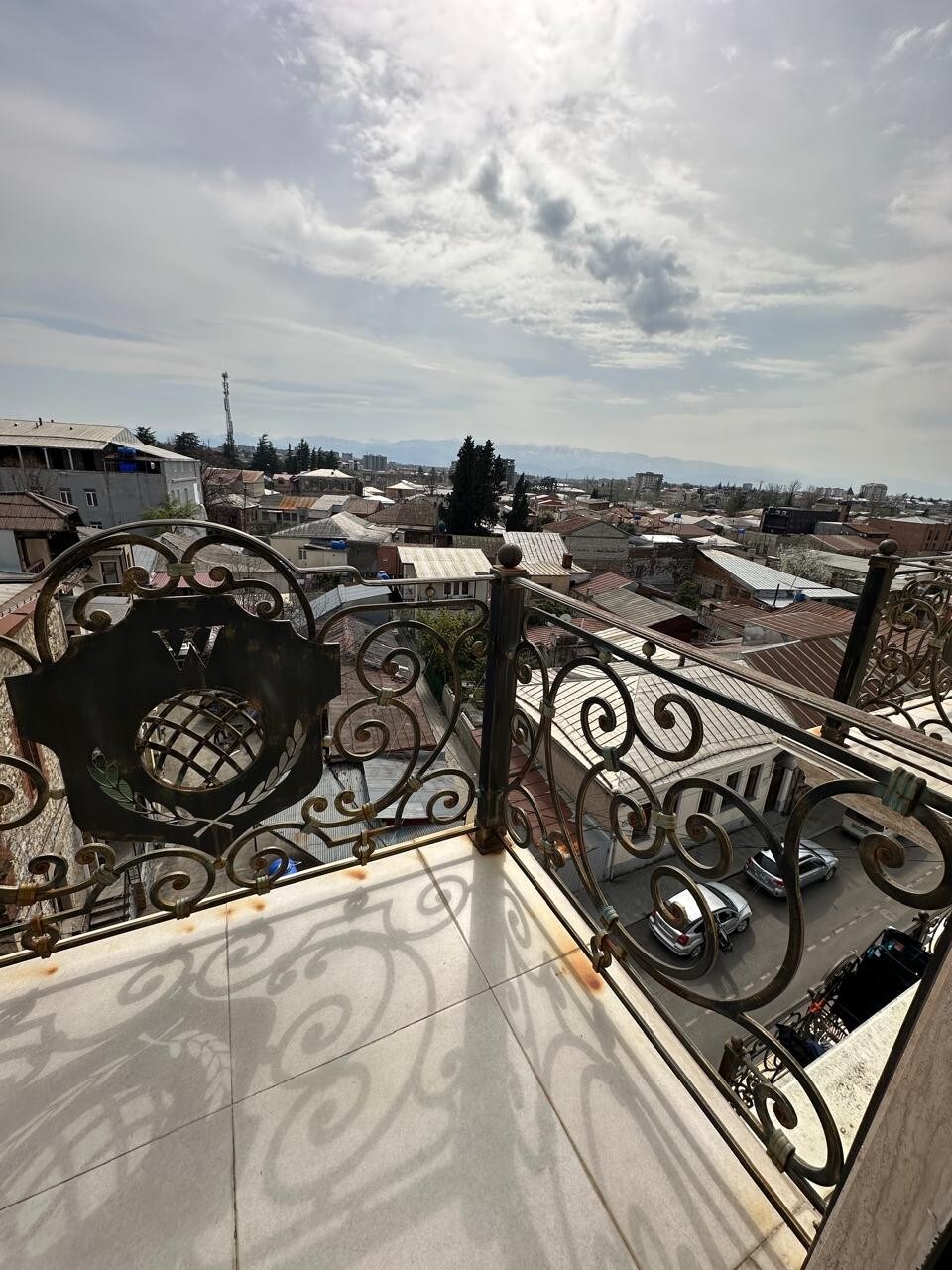 A balcony railing with intricate metalwork frames a view of the city. The rooftops of nearby buildings are visible under a cloudy sky, creating a sense of openness. The ground below shows a glimpse of vehicles along the street.