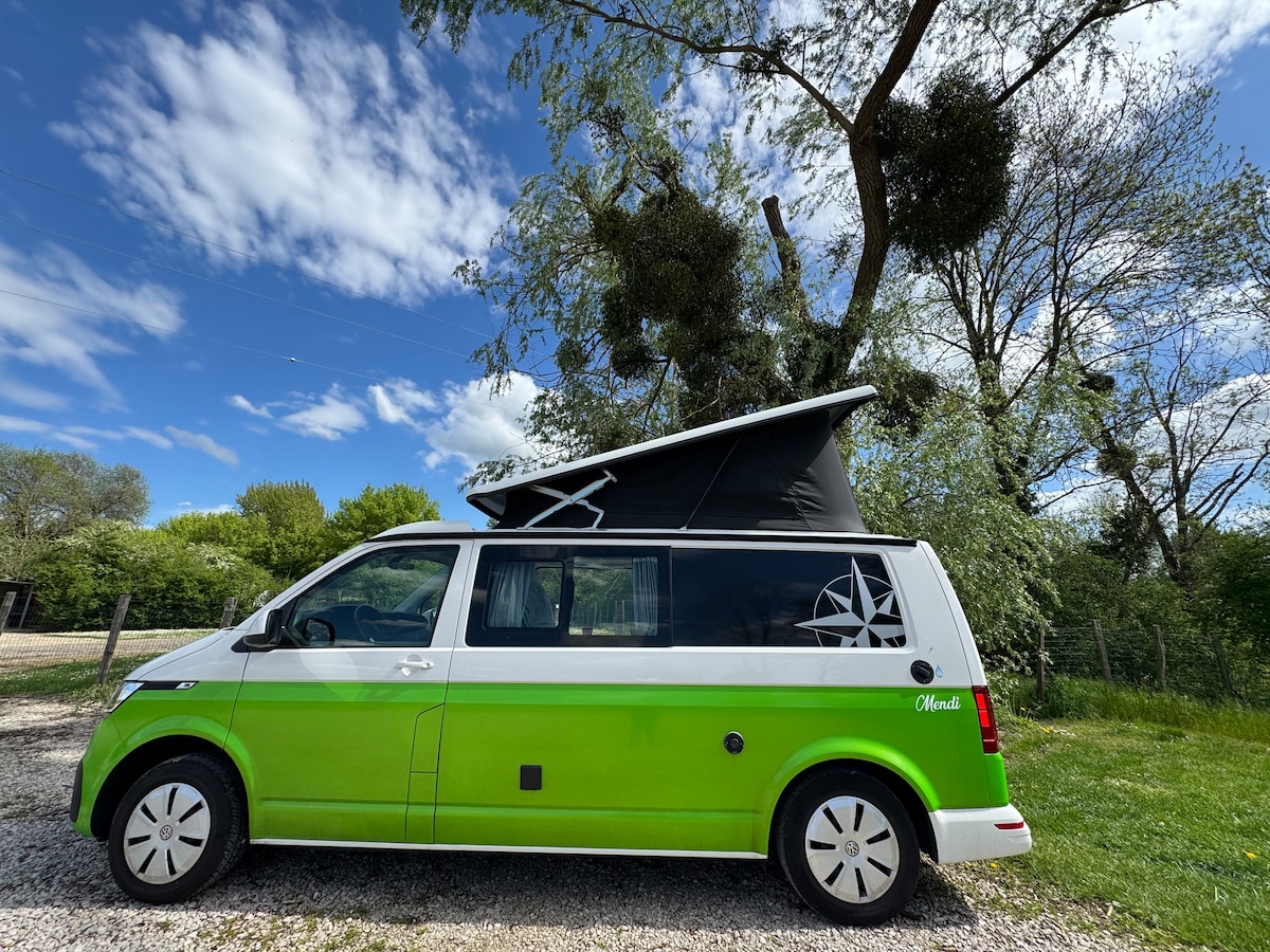 A Volkswagen Westfalia Kepler One camper van is parked on a gravel surface. Its vibrant green and white exterior features a pop-up roof. Surrounding greenery and a blue sky with scattered clouds create a natural backdrop. The van's design emphasizes adventure and mobility.