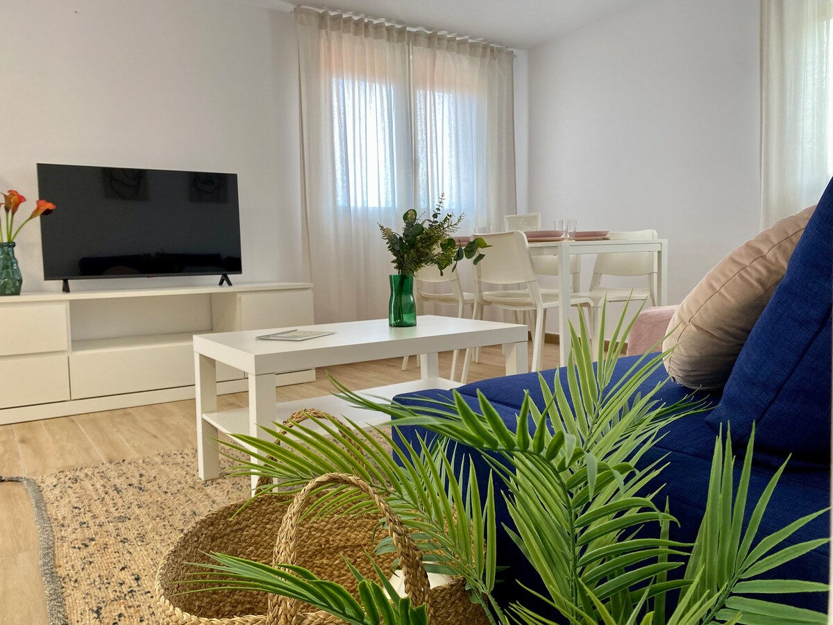 A bright living area features a blue sofa and a coffee table in front of a television. Natural light filters through sheer curtains, illuminating a dining space equipped with a white table and chairs. Decorative plants add a touch of freshness to the room.