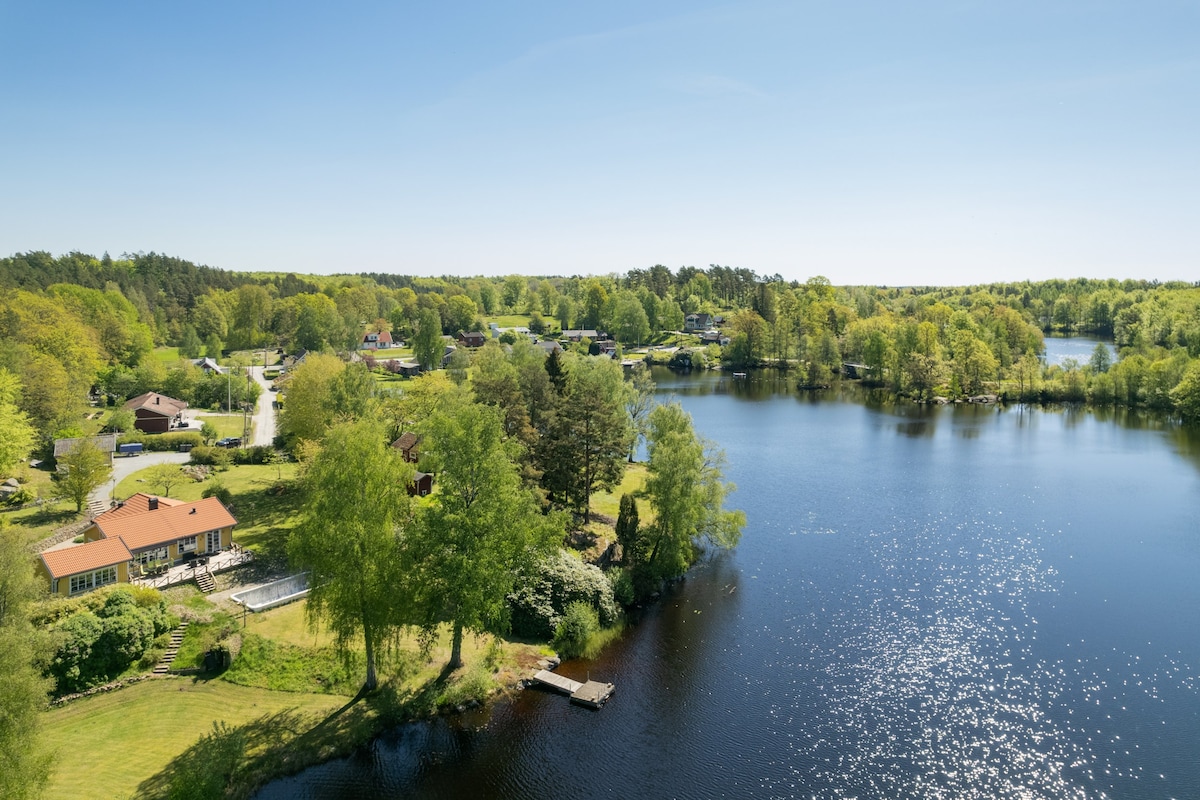 A serene landscape showcases a tranquil lake surrounded by lush greenery. The clear blue sky reflects on the water's surface, while several houses are nestled along the shoreline, enhancing the peaceful setting. A wooden dock extends into the lake, inviting exploration.