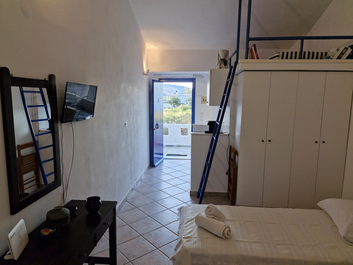 A simple interior space features a narrow sleeping area with a white bed and rolled towels. A wall-mounted television is visible near a glass door that opens to an outdoor area. Storage cabinets line one wall, and a mirror reflects the room's layout.