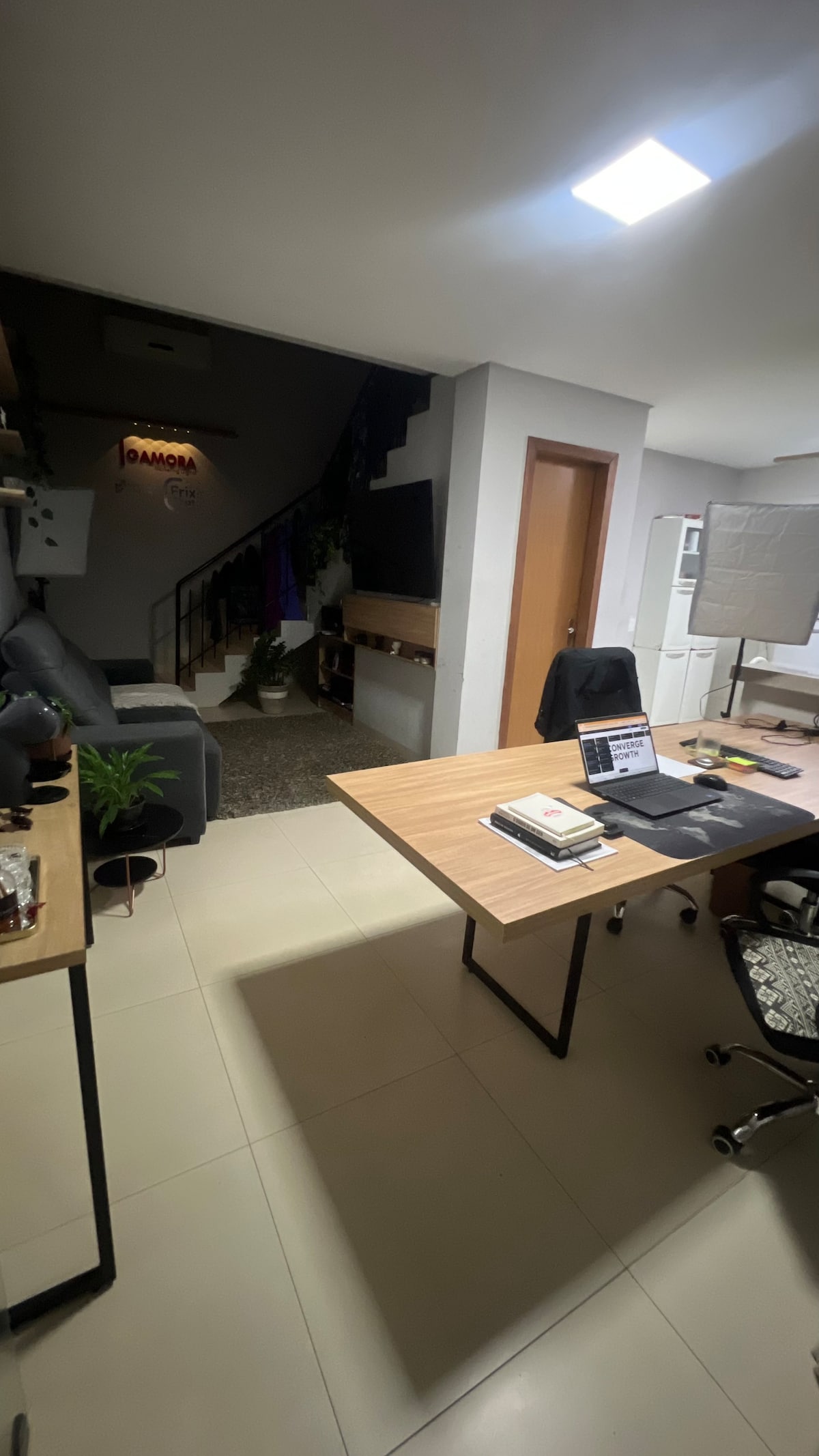 A modern workspace is depicted with a wooden desk positioned beside a comfortable chair. Shelving holds decorative plants and books, enhancing the functional layout. Soft lighting allows for a warm ambiance, while a staircase is visible in the background, leading to an upper level.
