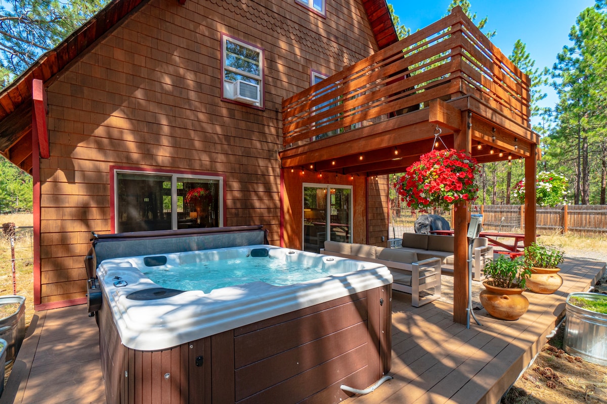 A spacious outdoor deck features a private hot tub and comfortable seating areas. Red accents from hanging flowers enhance the inviting space, and the cabin's rustic siding and large windows provide a warm backdrop against the surrounding forest.