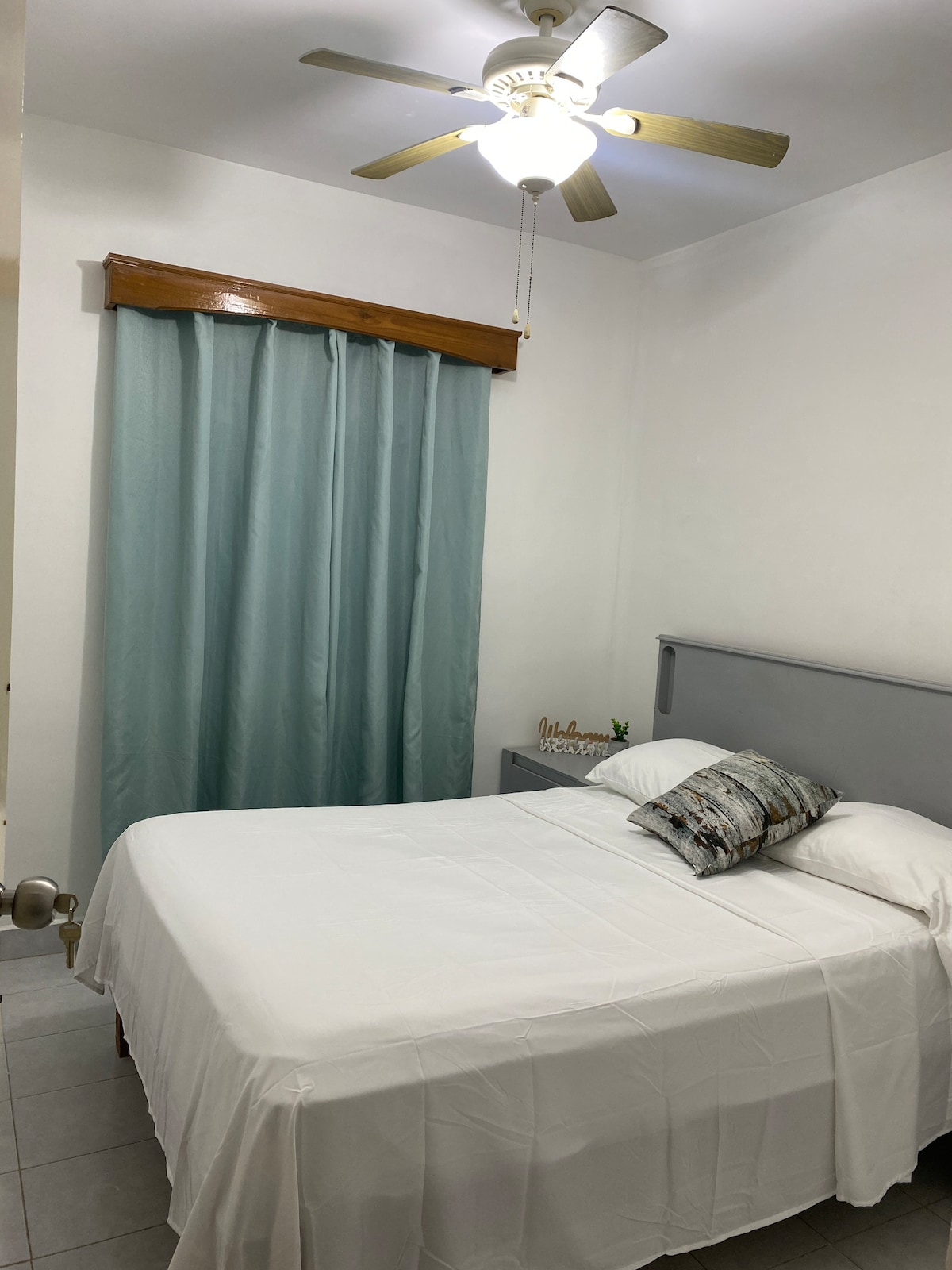 A neatly arranged sleeping area features a spacious bed with white linens, complemented by a decorative throw pillow. A soft blue curtain hangs beside the bed, and a ceiling fan provides airflow. A small nightstand displays a decorative piece, adding a personal touch.