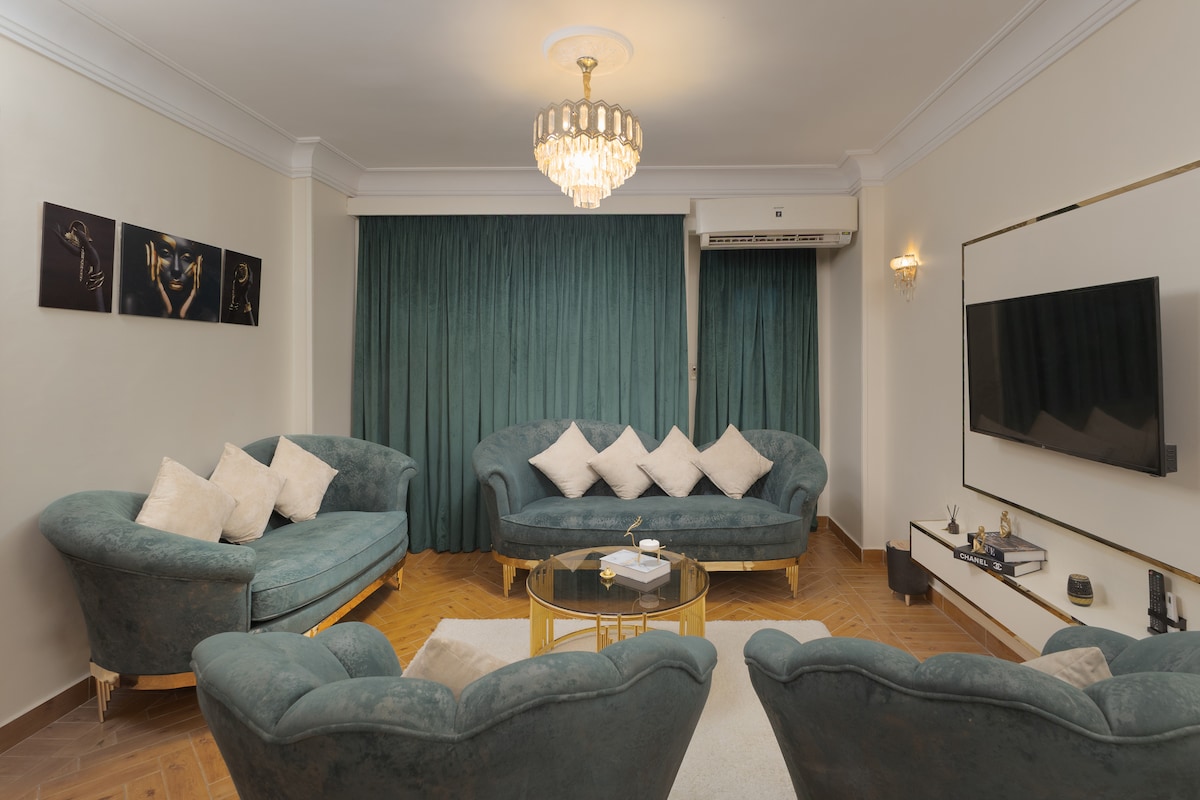A spacious living room features two plush sofas and a set of armchairs surrounding a circular coffee table. Large windows are dressed with curtains, allowing natural light to fill the area. An elegant chandelier hangs from the ceiling, enhancing the room's inviting ambiance.