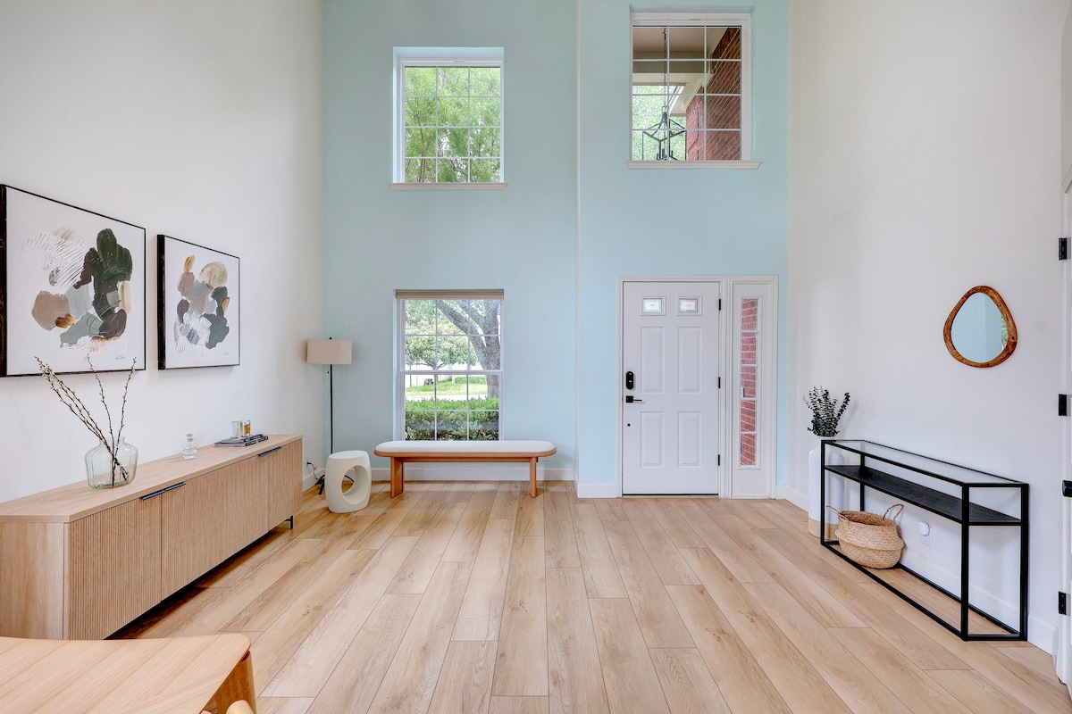 A bright entryway features tall ceilings and large windows that allow natural light to fill the space. A minimalist wooden bench is positioned near the entrance, accompanied by a sleek console table. Decorative wall art and a small plant add subtle touches to the inviting atmosphere.