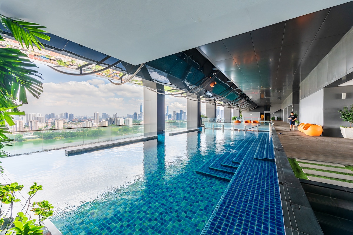 An infinity pool is depicted, surrounded by modern architecture with large windows revealing a skyline view. The water reflects the cityscape, while lounge chairs are seen nearby, providing a space for relaxation. Tropical plants add a touch of greenery to the area.