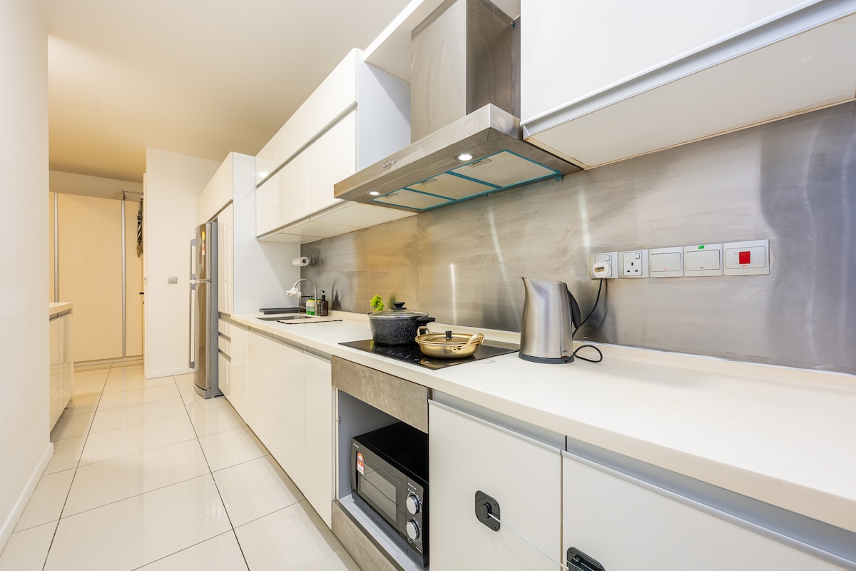 A modern kitchen is equipped with sleek white cabinetry and stainless steel appliances. A gray countertop features essential cooking items, including a kettle and a pot. The mirrored backsplash reflects light, while the spacious layout is complemented by tiled flooring.