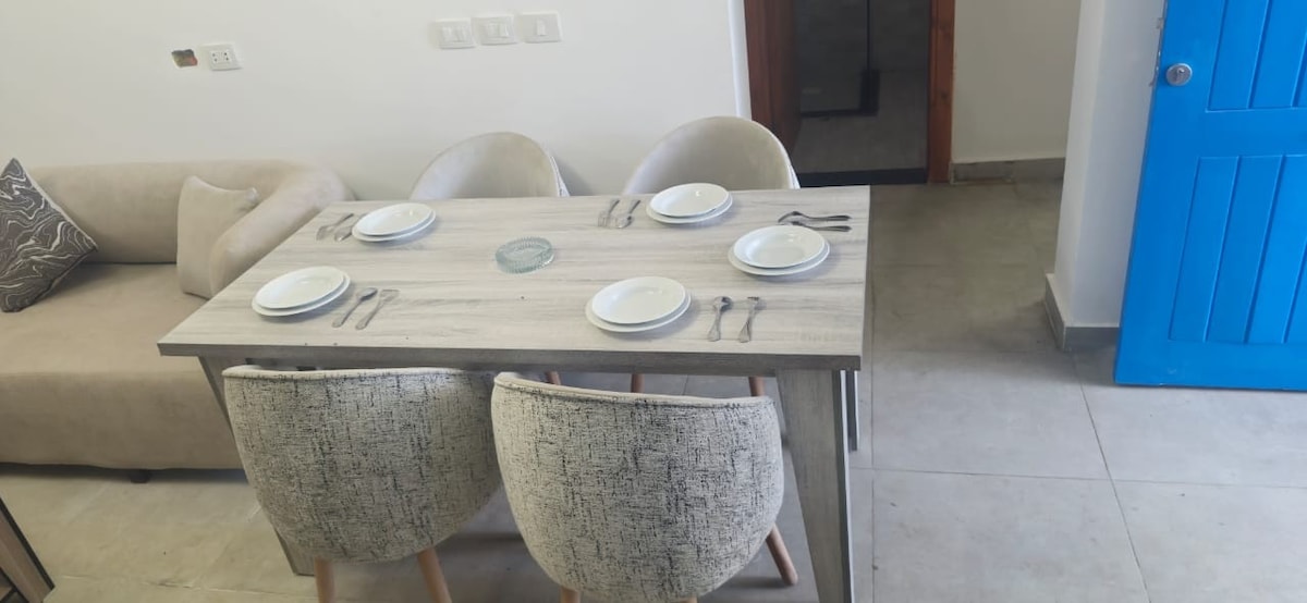 A dining table is set for four, featuring white plates and utensils, positioned near a soft, light-coloured sofa. The blue door adds a vibrant touch to the neutral tones of the room, which is illuminated by natural light.