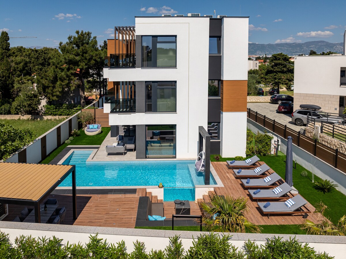 The modern villa presents a sleek exterior with large windows and a multi-level design. A private pool is bordered by a wooden deck with lounge chairs and shaded seating areas. Lush greenery surrounds the property, enhancing the outdoor space for relaxation.