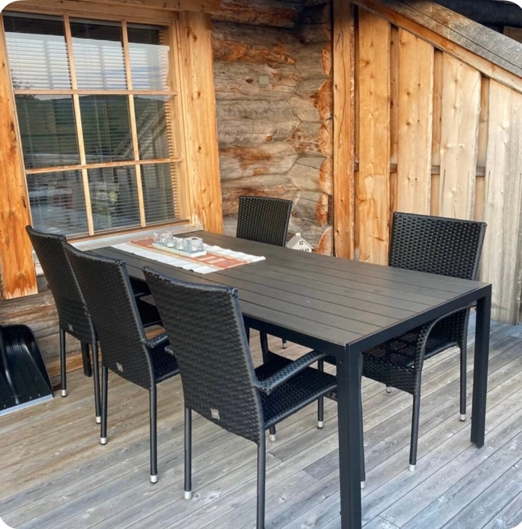 A spacious outdoor dining area is presented, featuring a rectangular table surrounded by six black wicker chairs. The rustic wooden backdrop complements the setting, and natural light streams through the nearby window.