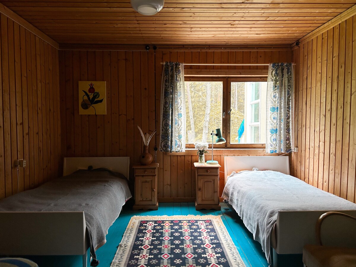 A warm, wooden room features two twin beds with light blankets. A colorful area rug with intricate patterns lies on the blue floor. Natural light enters through a window adorned with floral curtains, illuminating a bedside table with a lamp and decorative items.